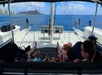Four people sit under shade at the stern of a catamaran sailing on blue water, with Waikiki skyline and Diamond Head crater visible in the distance under a partly cloudy sky.