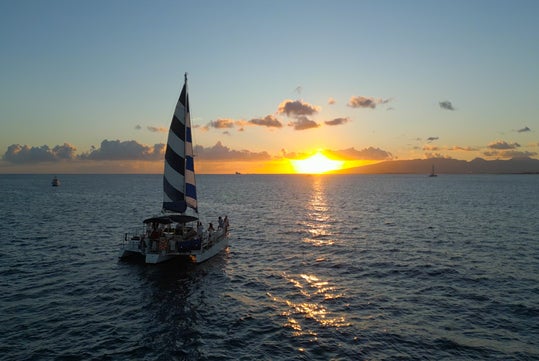 A sailboat with a blue and white striped sail is on the ocean at sunset, with the sun low on the horizon and scattered clouds in the sky.