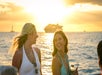Two women in swimsuits smile and talk by the ocean at sunset, with a large cruise ship visible in the background.
