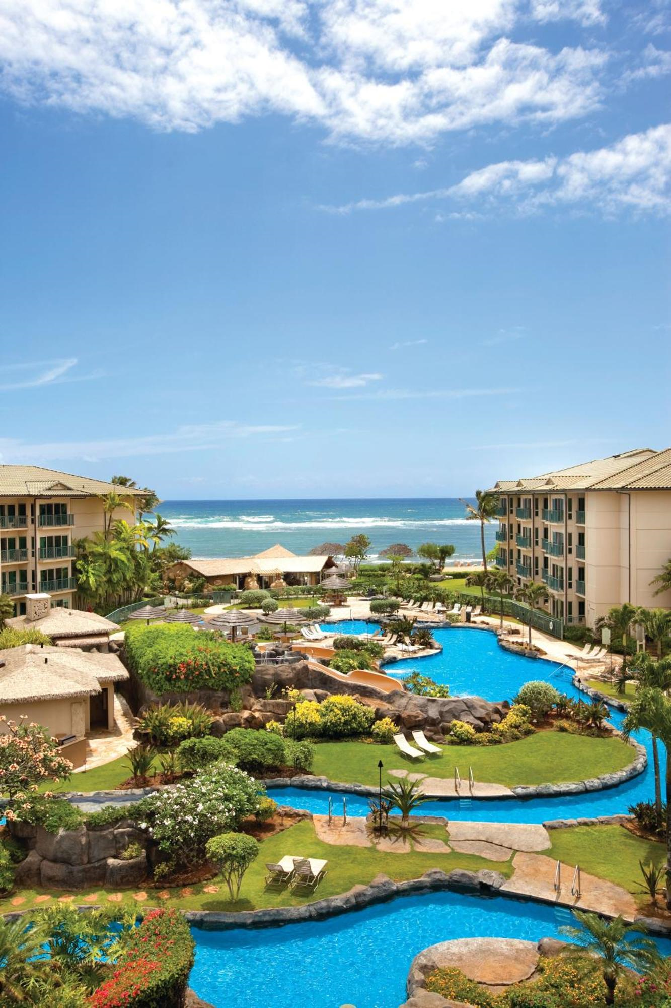 Aerial view of a resort with winding blue pools, lounge chairs, tropical landscaping, and two beige buildings facing the ocean under a partly cloudy sky.