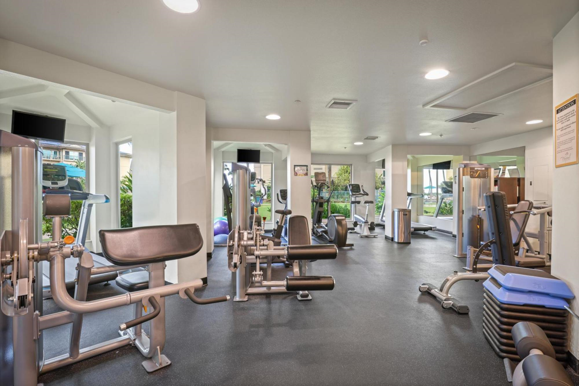 A gym room with various exercise machines, free weights, benches, and stacked aerobic steps. Large windows provide natural light and views of greenery outside.