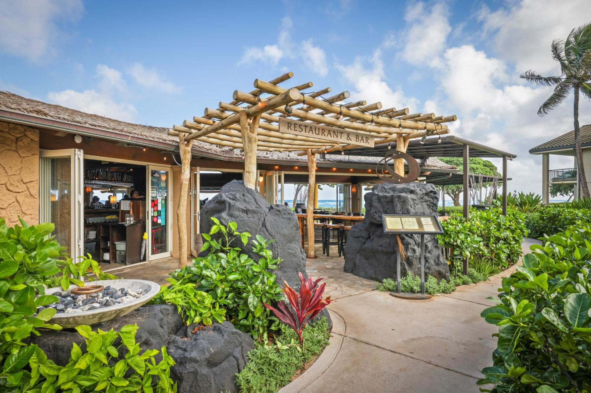 Outdoor restaurant and bar with wooden pergola, surrounded by rocks, plants, and ocean views under a partly cloudy sky.