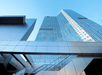 Modern skyscrapers with reflective glass facades rise against a clear blue sky, connected by an angular skybridge.