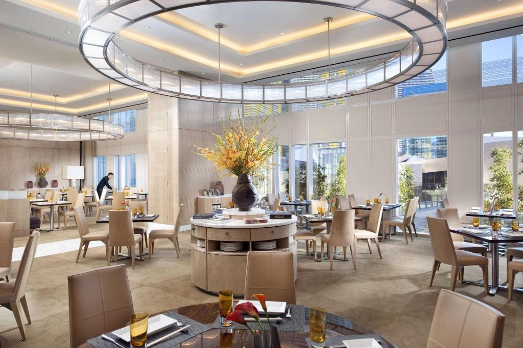 Elegant restaurant interior with round tables, beige chairs, large chandeliers, and floral centerpieces. A staff member is visible near the back. Large windows provide natural light.