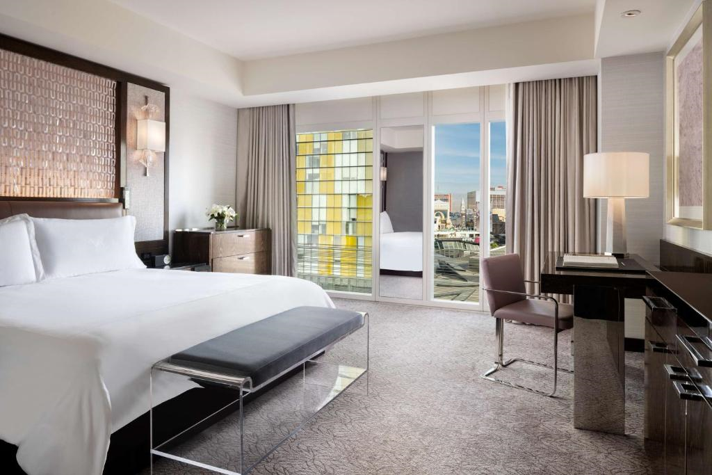 Modern hotel room with a large bed, desk, and glass doors opening to a balcony. Neutral tones, ample natural light, and cityscape views create a serene atmosphere.