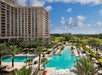 Large outdoor hotel pool surrounded by palm trees, lounge chairs, and umbrellas, with a multi-story hotel building and golf course in the background under a blue sky.