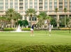 Two people play golf on a putting green in front of a large hotel, with palm trees and a fountain in the background.