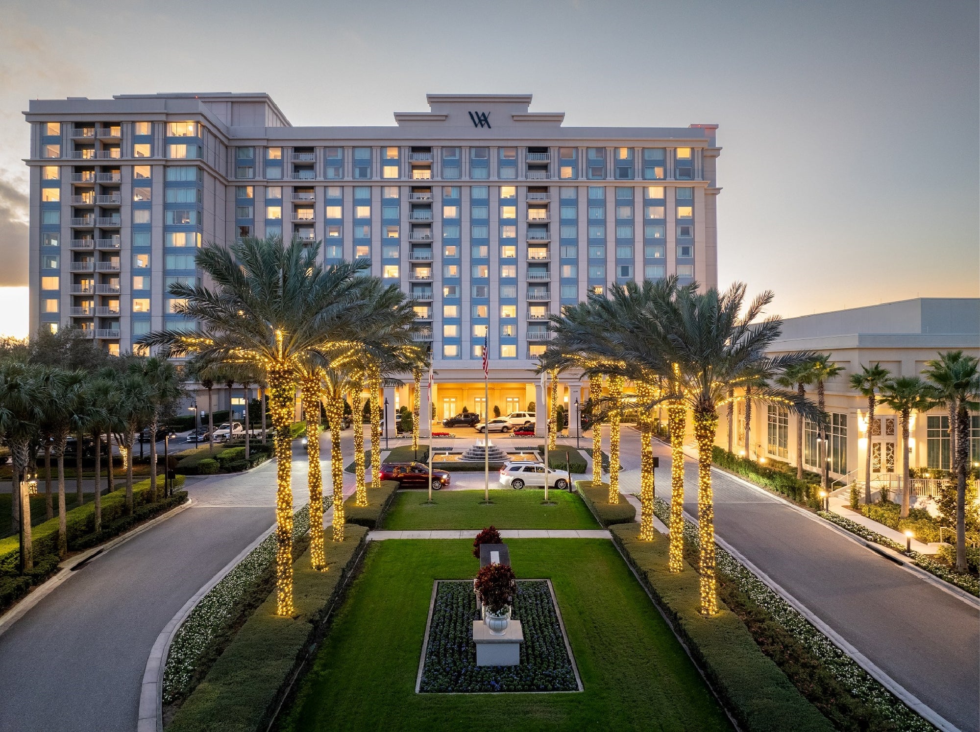 A large hotel with palm trees wrapped in string lights lines the entrance driveway at dusk, with cars parked near the building and a central statue in the landscaped median.