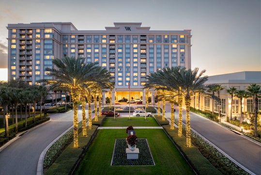 A large hotel with palm trees wrapped in string lights lines the entrance driveway at dusk, with cars parked near the building and a central statue in the landscaped median.
