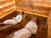 Two people in white robes lie on lounge chairs in a softly lit room with salt brick walls, appearing to relax.