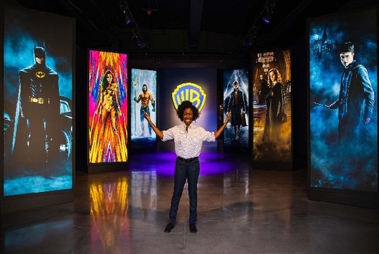A woman in a white shirt and dark pants smiling with her hands up and out with several movie posters and the Warner Bros. logo behind her.