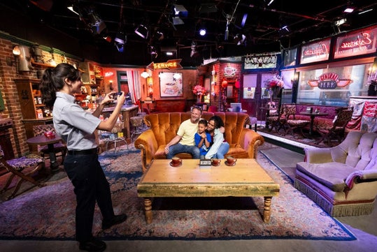 A family poses for a photo on an orange couch in a replica of a coffee shop set, while a woman takes their picture. Bright lights and props decorate the background.