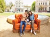 A family of three sitting on an orange couch taking a selfie with a directly fountain behind them and a bus and a couple of building in the background.