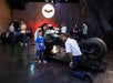 People interact with a Batcycle display in a museum-like setting, with a large illuminated Batman logo on the wall in the background.