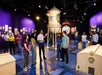 A group of people listens to a guide inside a Warner Bros. Studios exhibit featuring a large model water tower and animation displays.