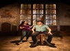Two people in a room with stained glass windows; one sits on a stool wearing a pointed hat, while the other stands beside, gesturing and smiling.
