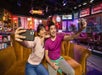 Two people smiling and taking selfies while sitting on a yellow couch with a TV show set behind them on the Warner Bros. Studio Tour Hollywood.