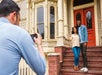 A couple posing on brick steps leading up to a cream colored house with a red door with a man at the bottom of the stair taking their photo.