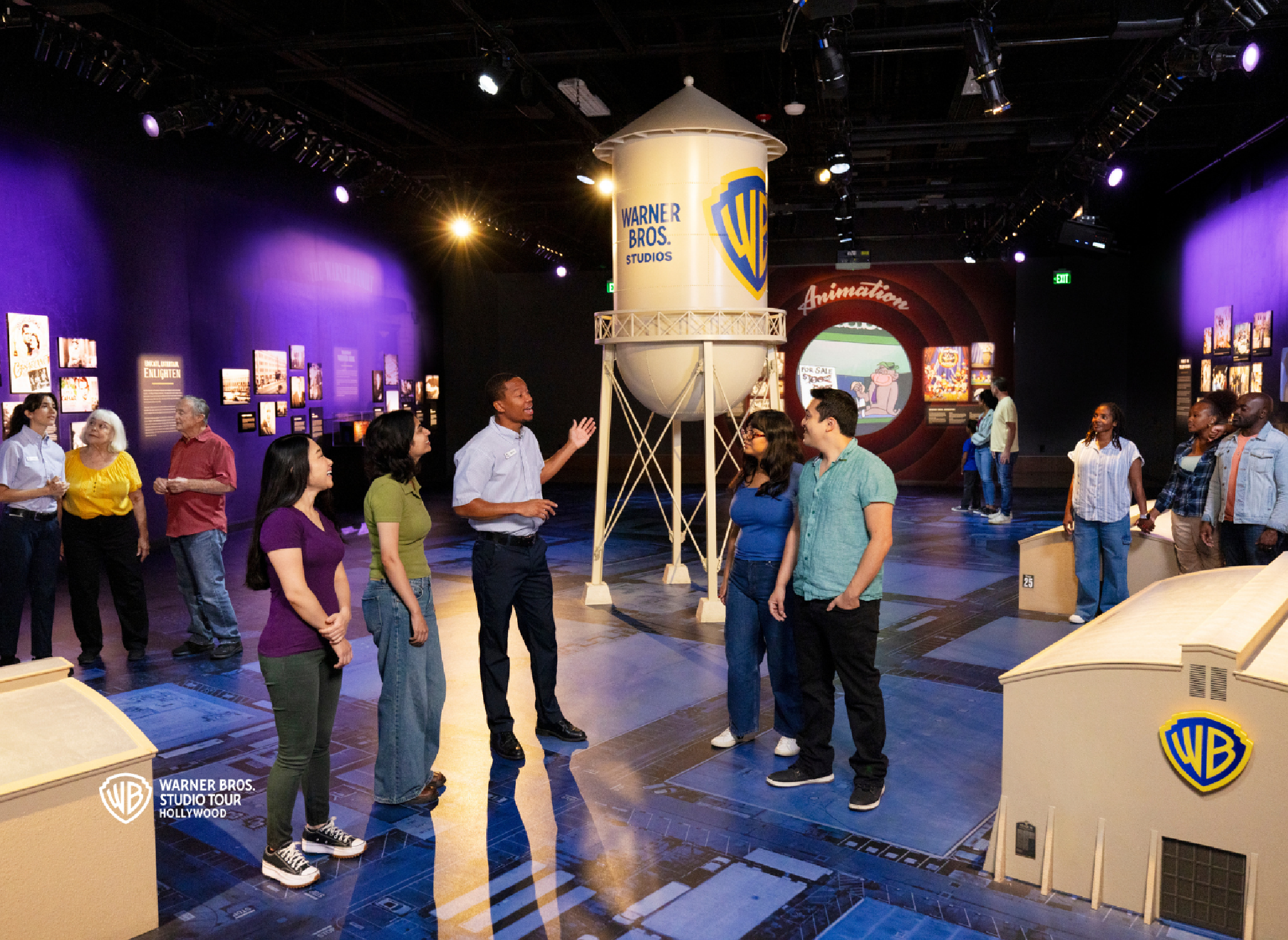 A group of people stand and listen to a guide near a large Warner Bros. water tower replica inside an indoor studio tour exhibit.