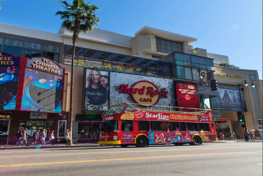 A red StarLine CitySightseeing tour bus drives past the Hard Rock Cafe on a sunny street lined with palm trees and colorful signs.