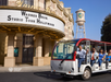 A Warner Bros. Studio Tour tram with visitors passes in front of the studio entrance and water tower in Hollywood on a sunny day.