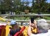 People on a double-decker tour bus take photos of a yellow Beverly Hills sign on a sunny day, surrounded by trees and a park area.