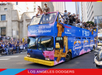A double-decker bus decorated with "2024 World Champions" and Los Angeles Dodgers logos drives through a city street during a victory parade with cheering fans on the sidewalk.