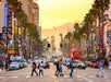 People crossing a busy street in Hollywood at sunset, with palm trees lining the road and various billboards and signs on buildings. Traffic fills the intersection.