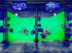 Two people pose on a green screen set. One simulates flying on a broomstick, and the other rides a motorcycle. Various cameras and monitors surround them.