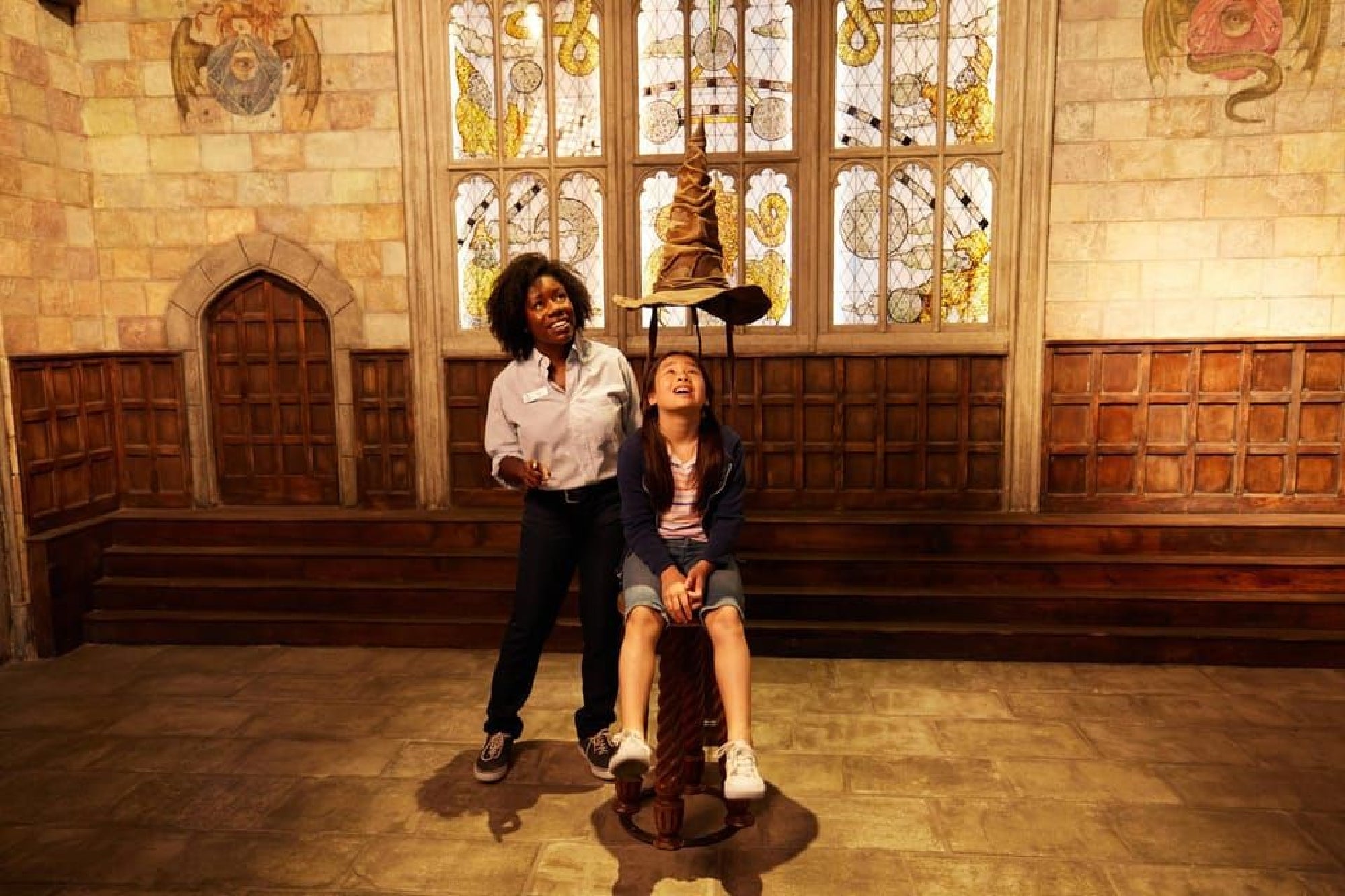 Person seated wearing a pointed hat, smiling, with another person standing nearby in a room with stone walls and stained glass windows.
