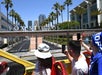 Tourists on an open-top bus take photos of the Fox Studios entrance, surrounded by palm trees and adjacent modern buildings, with skyscrapers visible in the background.