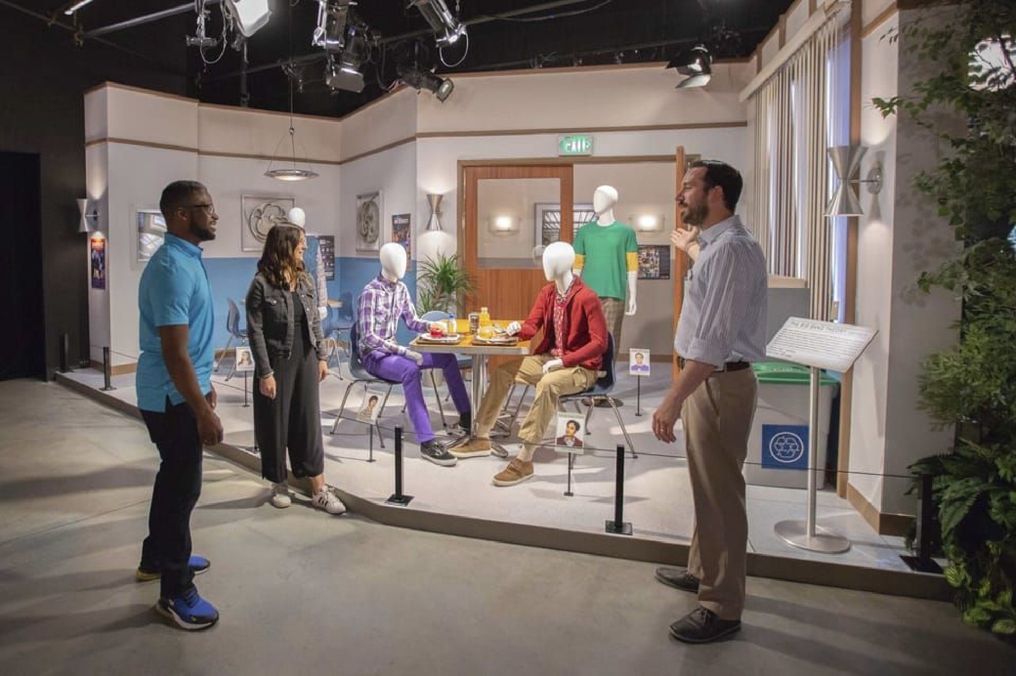 Four mannequins seated at a table in a display resembling an office break room, with two people observing. The scene includes props like food items and a recycling bin nearby.