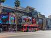 A red tour bus passes by the Hard Rock Cafe and TCL Chinese Theatres on a sunny street lined with palm trees.