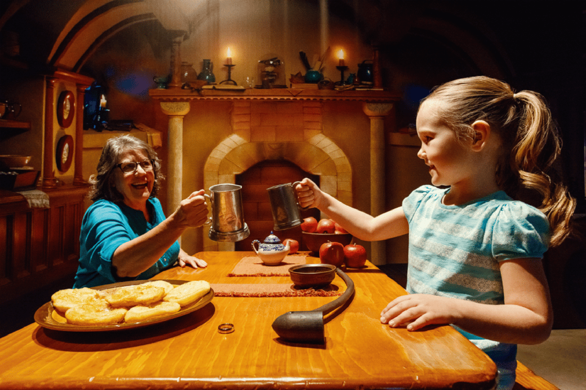 A woman and a girl toast with metal mugs at a wooden table set with food in a warmly lit room.