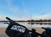 A bicycle with a bag labeled "Unlimited Biking" is parked near a body of water, with the Washington Monument visible in the background under a clear sky.