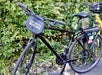 A black bicycle with a front bag labeled "Unlimited Biking" is parked on a paved path next to green foliage.