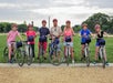A group of six people wearing helmets stand with their bicycles on a grassy area, with trees and monuments visible in the background.