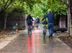 A group of people wearing helmets walk and ride bicycles on a damp, tree-lined path. The ground reflects red light, and there are fallen leaves scattered around.