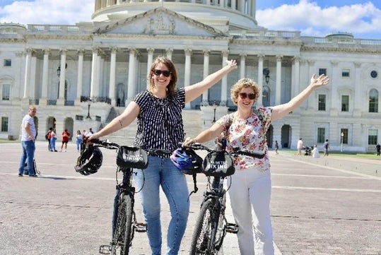 Two women stand with bicycles in front of a large government building, raising their arms and smiling; bike helmets rest on the handlebars.