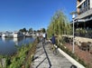 Two people ride bikes along a wooden waterfront path beside a marina with docked boats and a restaurant patio on a sunny day.