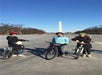 Three people wearing helmets stand with bicycles on a paved area in front of the Washington Monument under a clear blue sky.