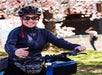 A woman wearing a helmet and sunglasses smiles while standing with her bicycle near blooming cherry blossoms.