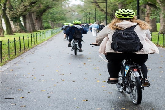 People riding bicycles on a tree-lined path, wearing bright green helmets and backpacks.