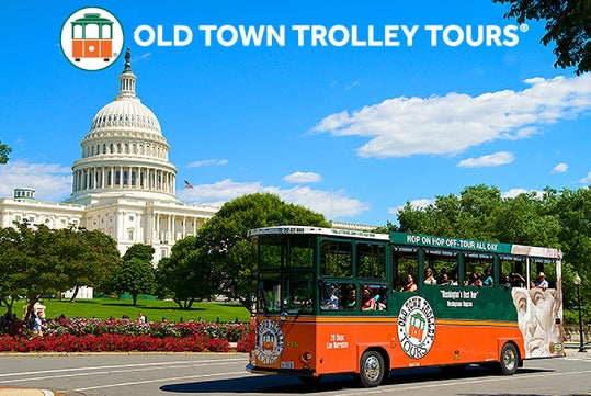 A green and orange Old Town Trolley Tours bus drives past the U.S. Capitol building on a sunny day, with trees and flowers in the background.