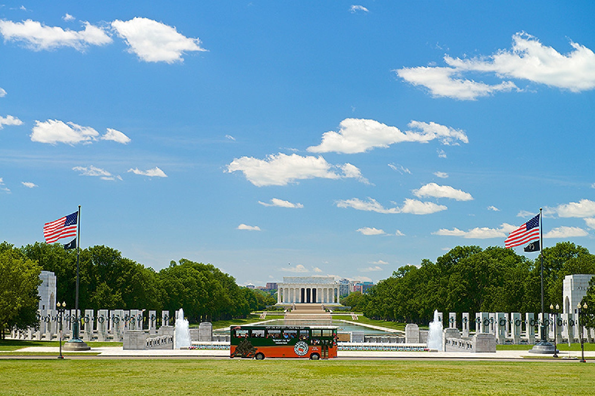 A sightseeing trolley is parked in front of the National World War II Memorial, with the Lincoln Memorial and two American flags in the background under a blue sky.