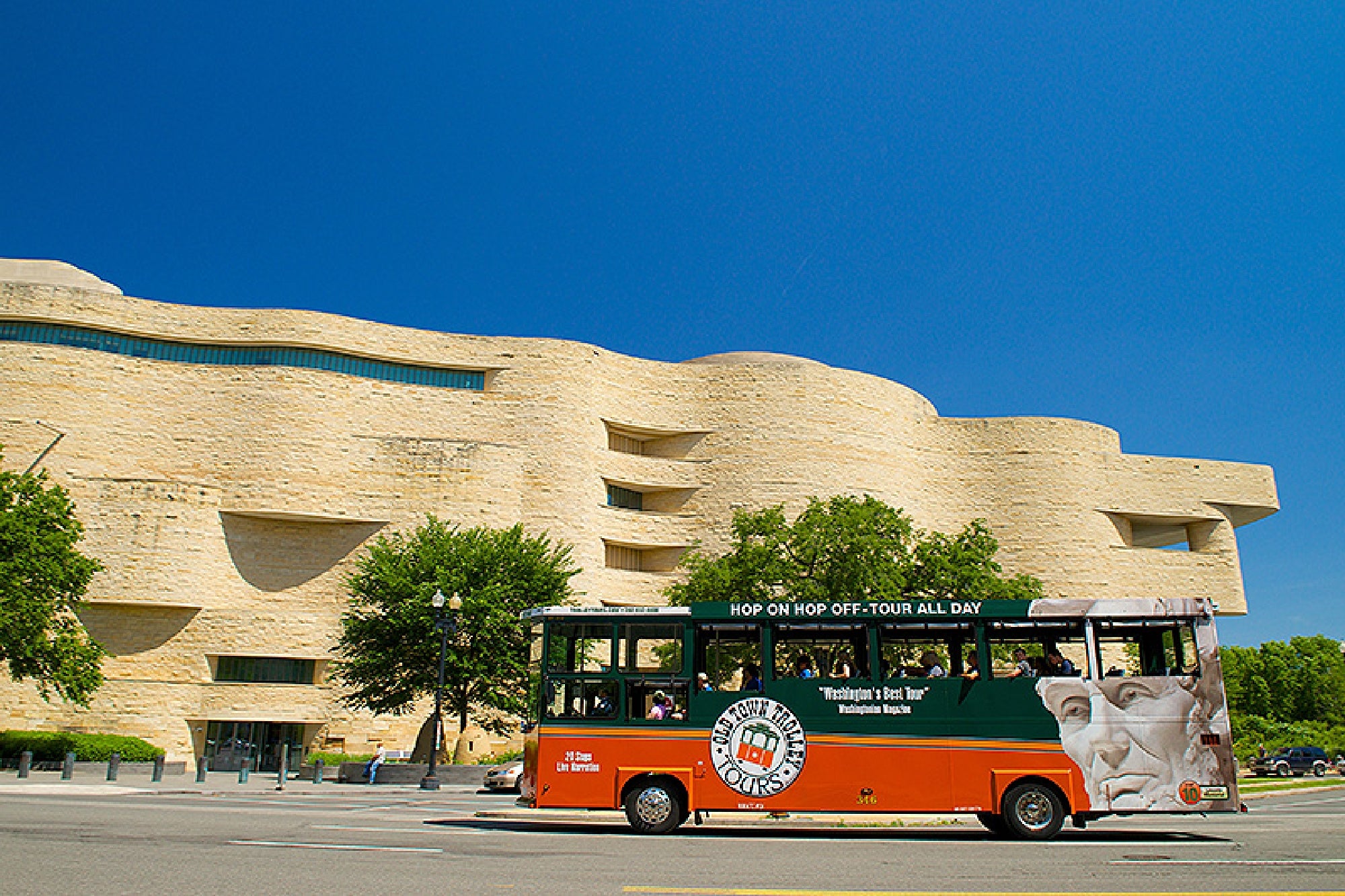 A green and orange open-air tour bus drives past the National Museum of the American Indian under a clear blue sky.