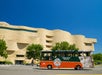 A green and orange open-air tour bus drives past the National Museum of the American Indian under a clear blue sky.