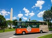A CitySights Washington DC tour bus drives near the Washington Monument on a sunny day with a blue sky and scattered clouds.