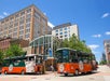 Two orange and green tour trolleys are parked on a city street in front of a multi-story building on a sunny day.