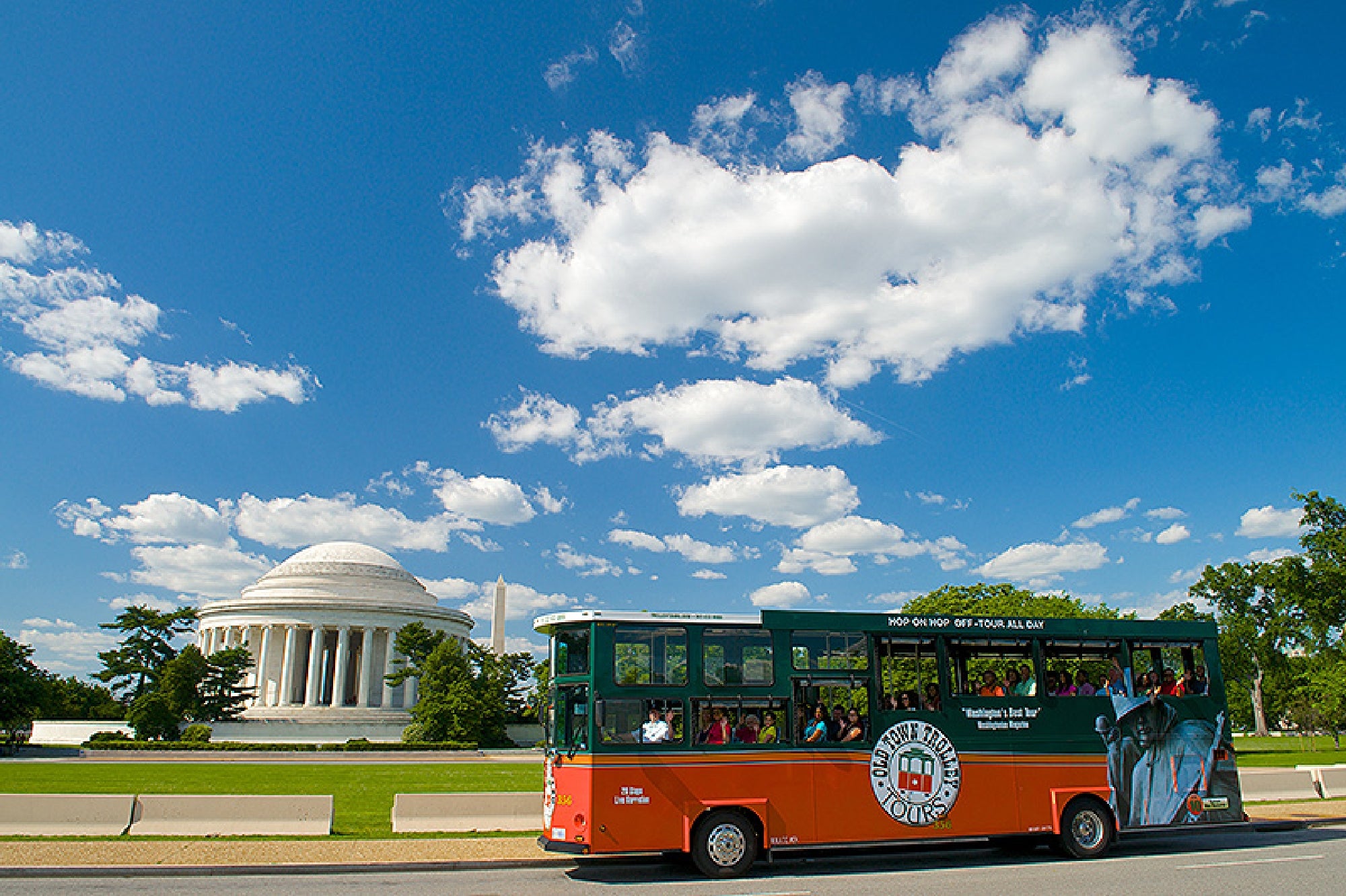A tour bus drives past the Jefferson Memorial under a blue sky with scattered clouds in Washington, D.C.